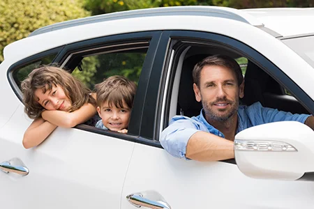 A man and two children smiling through the open windows of a parked white car